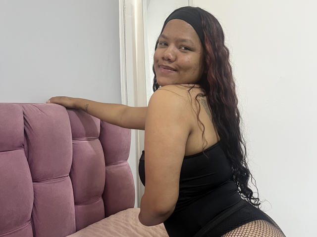 ScarlettNoir with long red-black hair, black headband, black dress, fishnet stockings, smiling, leaning on pink velvet chair, showing her back and small tattoo on right arm.