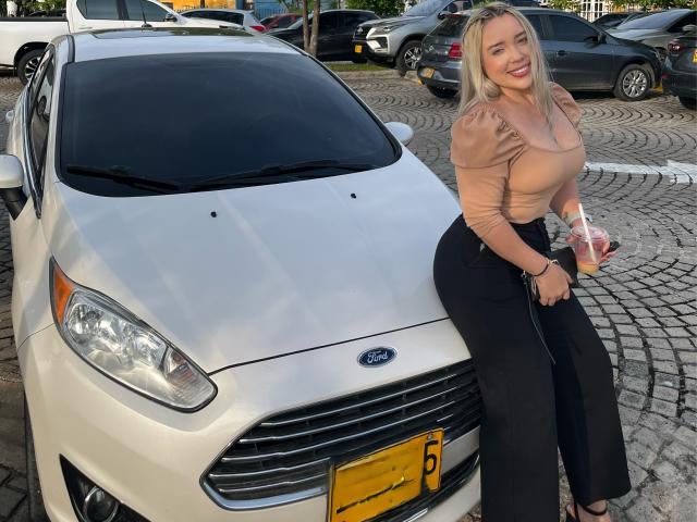 MiaBlondeHot with big tits, blonde hair, and a smile, sitting on a white Ford car, wearing a tan top and black pants, holding a drink.