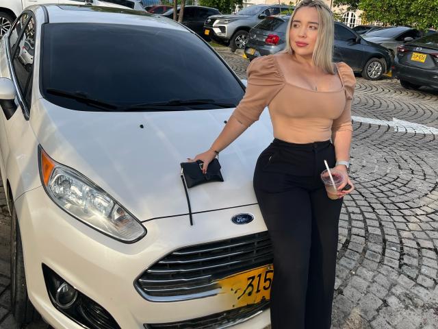 MiaBlondeHot with blonde hair, tan top, black pants, holding drink, leaning on white Ford, large boobs, black phone, yellow license, curvy, standing in parking lot, daytime.