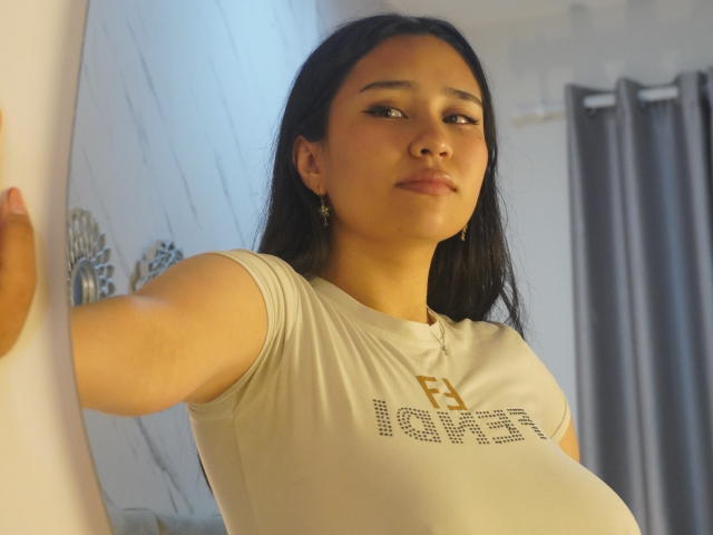 Karollteyes with long black hair, tan skin, and small gold earrings, wearing a white shirt that shows her big tits, standing with arms out, looking slightly to the side.