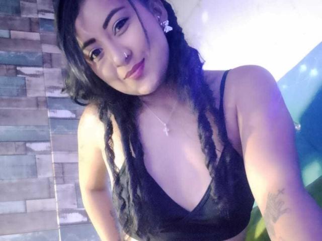 SexySerenaHot with black dreadlocks, nose ring, and cross necklace, wearing a black lace bra, smiling, showing her tits, tattoo on her right arm, in a dimly lit room.