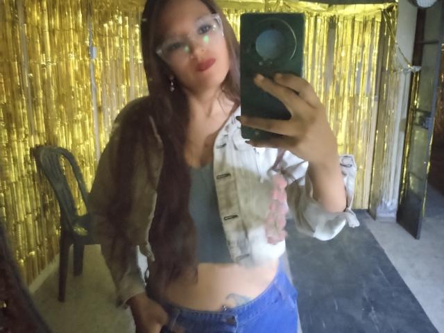 UrsulaHot69 with long brown hair, wearing glasses, a grey crop top, blue jeans, and a white jacket, holding a green phone, showing a flaccid pink dick in mirror selfie.