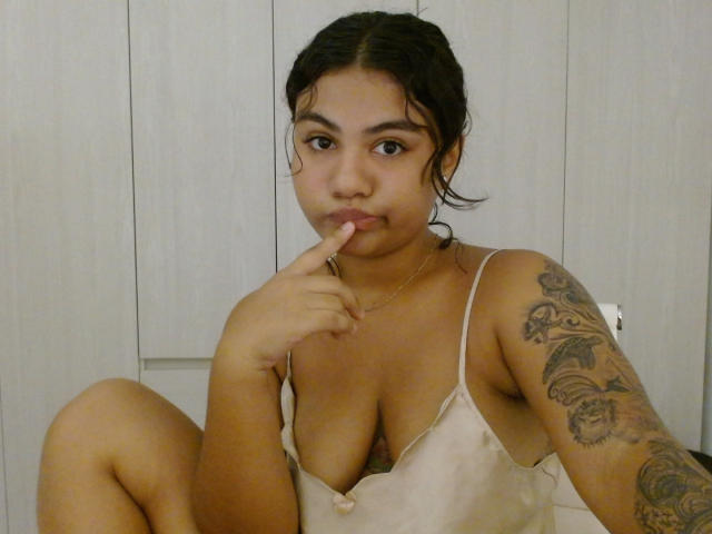ParadiseBloom, con gli occhi marroni e i capelli scuri, indossa una canotta bianca, si succhia il dito, mostra il seno e ha un braccio tatuato, seduta con le ginocchia sollevate..