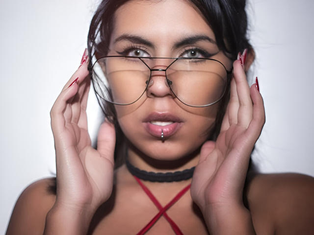 Dominalia mit Brille, Hände im Gesicht, rote Nägel, schwarzer Choker, rotes Oberteil mit Kreuzmuster, durchstochener Mund, Blick in die Kamera, braune Augen, dunkles Haar, weißer Hintergrund, leicht geöffneter Mund..