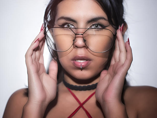 Dominalia mit Brille, Händen im Gesicht, roten Nägeln, einem Choker und einem gestochenen Mund, die ihre Brüste und ihren Hintern mit einem sexy, selbstbewussten Blick und einem leichten Grinsen zur Schau stellt..