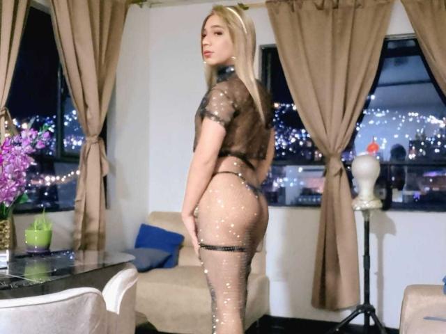 Cataline, cheveux blonds, vêtue d'une tenue noire en résille transparente, montrant ses fesses et sa vulve, se tient dans une pièce avec des rideaux beiges et une vue sur la ville la nuit..