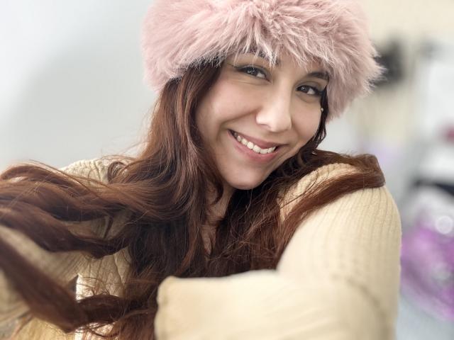 AmberlStha with long brown hair, pink fluffy hat, smiling, wearing a beige ribbed sweater, her tits slightly visible under the sweater, in a selfie with blurred background.