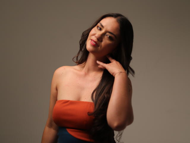 TheresaLarson with long brown hair, wearing an orange strapless top, slightly smiling, hand touching her neck, showing her tits, against a plain gray background.