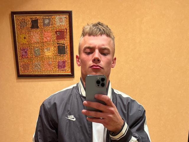 EthanWilder with short blonde hair, wearing a gray Adidas jacket, taking a selfie with a silver iPhone in a beige-walled room, looking slightly puffy-eyed.