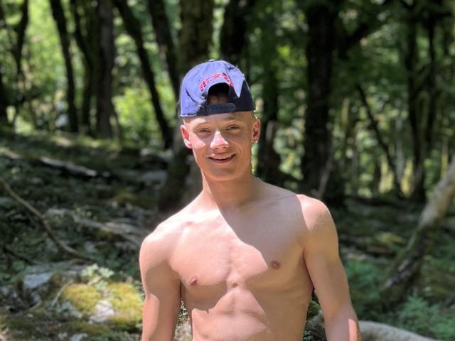 Shirtless EthanWilder with a blue baseball cap, smiling, standing in a forest, showing a muscular chest and a small, flaccid, uncircumcised penis.
