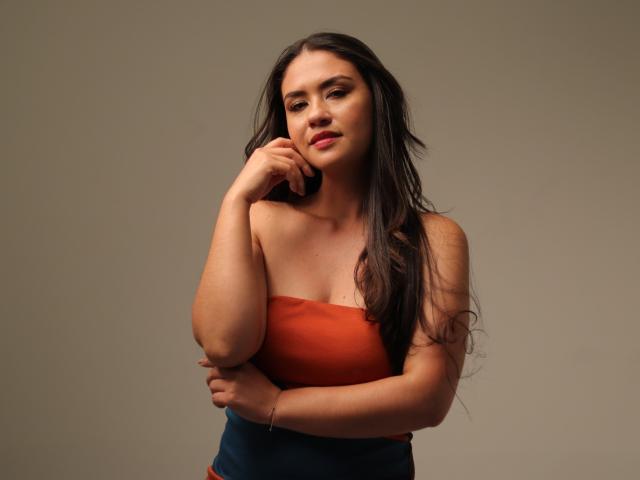 TheresaLarson with long dark hair, wearing an orange strapless top, has her hand near her mouth, looking slightly to the side with a subtle smile, showing her tits.