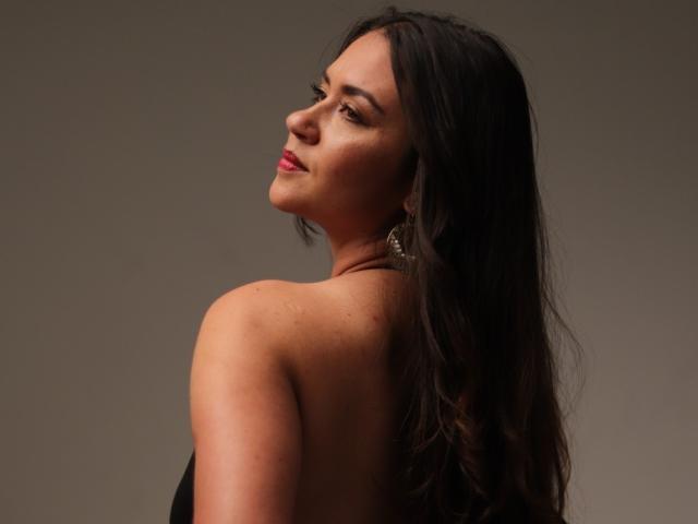 TheresaLarson with long black hair, looking over her shoulder, showing her bare back and small, round ass, wearing a black top and gold earrings, with a slight smile.