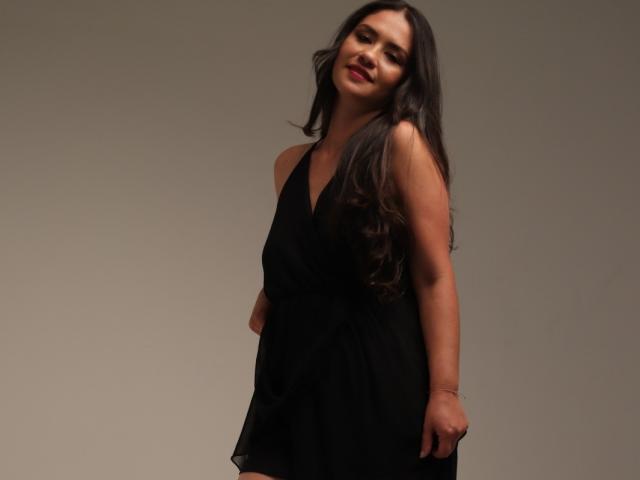 TheresaLarson with long dark hair, wearing a black halter dress, smiling slightly, standing with hands in dress, bare shoulders, brown skin, subtle makeup, and red lipstick.