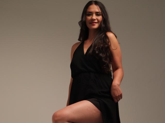 TheresaLarson with long dark hair, wearing a black sleeveless dress, has her right leg raised, showing her thigh, smiling with red lips, standing against a plain gray background.