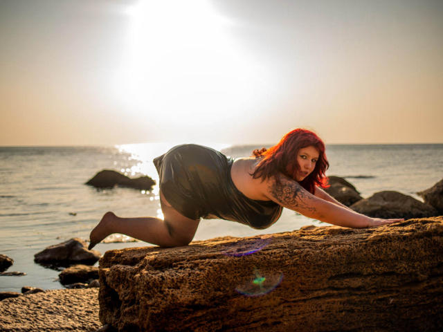 LiliyaMoon, con i capelli rossi, è accovacciata su una roccia, indossa un vestito nero che mette in mostra il suo braccio tatuato e i suoi fianchi sinuosi, su una spiaggia rocciosa illuminata dal sole, e guarda verso la fotocamera..