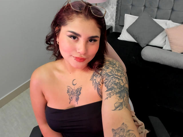 KatiaRodriguez with red curly hair, wearing a black strapless top, showing tattoos on her right arm and chest, and a butterfly tattoo above her tits, sitting in a bedroom.