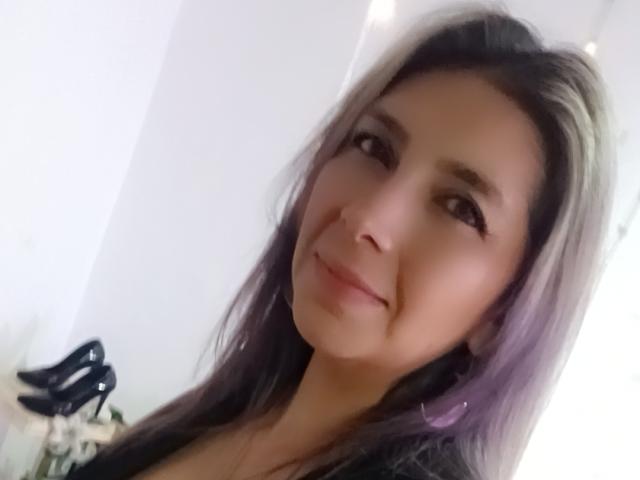 MichelleDumont with long dark hair and purple highlights, smiling slightly, brown eyes, light skin, wearing a black top, black high heels in the background, soft lighting.