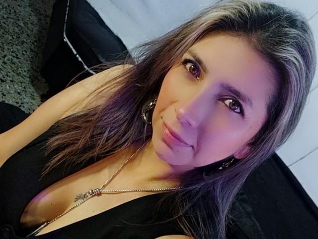 MichelleDumont ha gli occhi marroni, i capelli lunghi castani e un naso piccolo. Indossa una maglia nera con un taglio basso, che lascia intravedere un po' il seno. Sta sorridendo leggermente..
