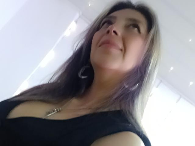 MichelleDumont with a slight smile, looking up, has medium-length brown hair, black top, and a necklace, showing her tits. Low-angle selfie, soft lighting.