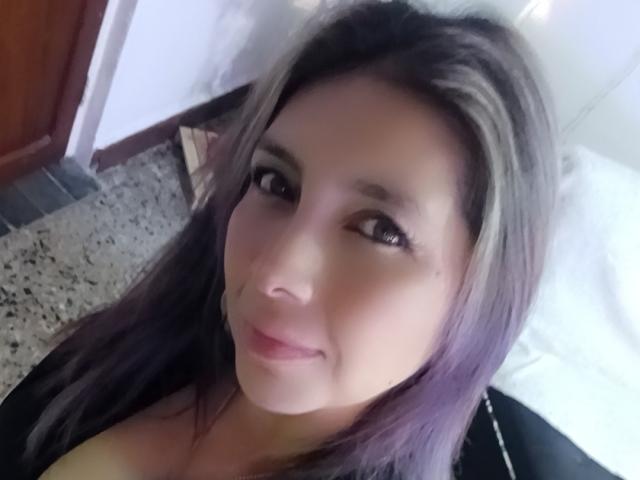 Il viso di MichelleDumont, con la sua pelle marrone chiaro, i capelli viola e marroni, gli occhi marroni e un piccolo sorriso. Le sue labbra rosa sono leggermente dischiuse. Indossa una maglietta nera..
