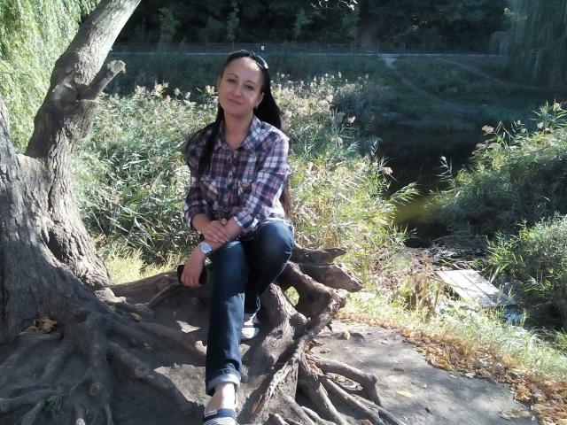 MelissaShell with medium tits, long black hair in braids, wearing a plaid shirt, jeans, and sandals, squatting on a tree root, smiling, in a sunlit, wooded area.