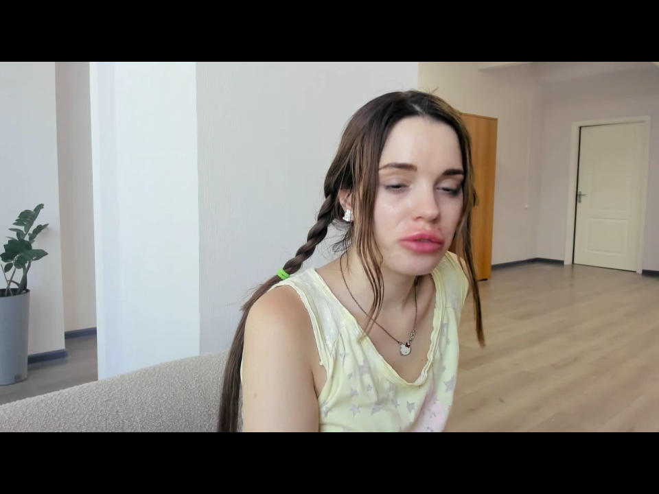 VeraWow with puffy red lips, long brown braid, white sleeveless top, small tits, green earbuds, necklace, sitting on couch, wooden floor, white walls, door in background.