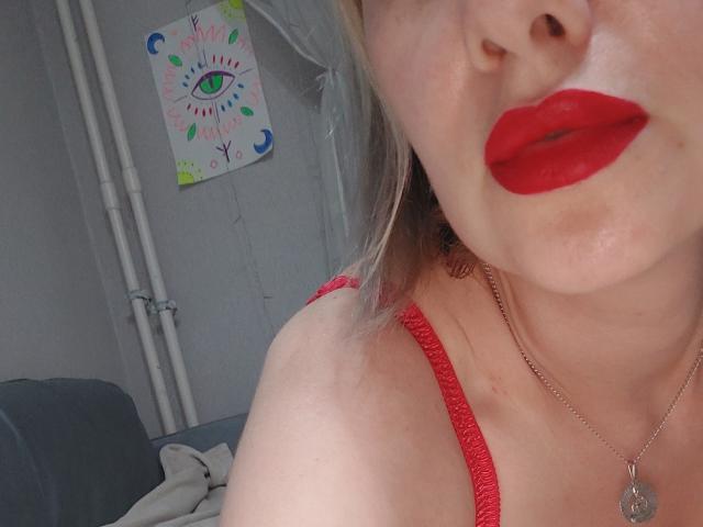 Il viso di BlueOli con rossetto rosso, labbra leggermente serrate, una spalla visibile con una maglietta a spalline rossa, una collana d'argento con un ciondolo a forma di cuore, capelli biondi, pelle chiara, una parete bianca sullo sfondo..