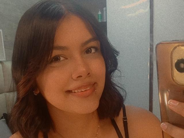 EmilyRoberts has medium brown skin, shoulder-length wavy black hair, a lip piercing, and is smiling at a mirror selfie with a gold phone, wearing a black strap top.