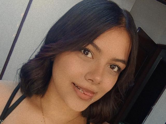 EmilyRoberts has medium brown skin, dark brown wavy hair, brown eyes, and a subtle smile with a small nose piercing. She's wearing a black strap top and a gold necklace.