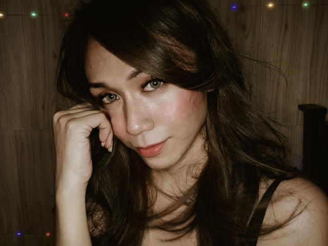TsJade has wavy brown hair, light skin, and a subtle smile with her hand resting on her cheek. She's wearing a black top, looking cute and sexy.