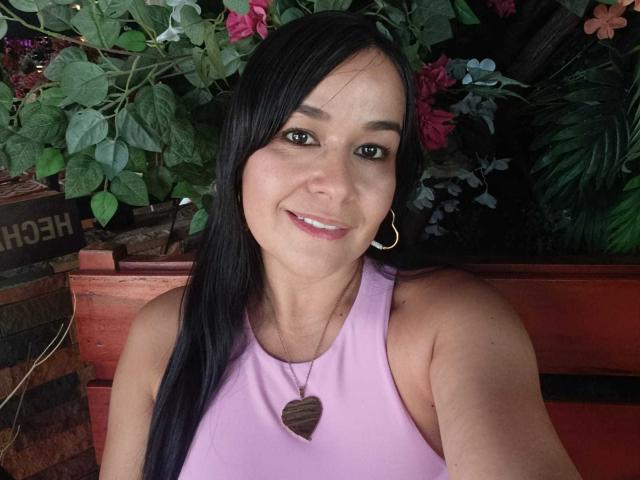 TamaraFoxy with long black hair, smiling, wearing a pink sleeveless top, gold hoop earrings, and a heart-shaped necklace, sitting on a red wooden bench.