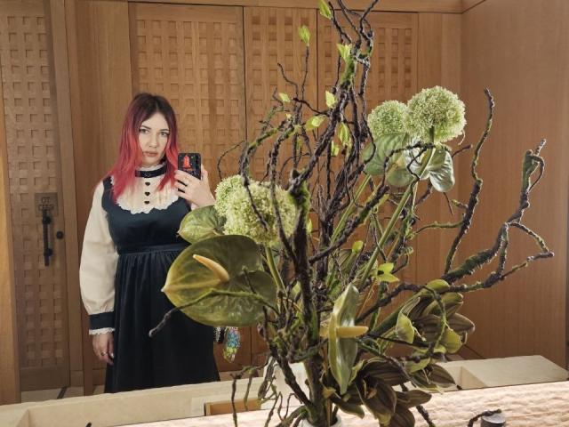 BlueOli mit rot gefärbten Haaren, trägt ein schwarz-weißes Kleid, hält ein Telefon und steht neben einer Blumenanordnung mit grünen Blättern und weißen Blumen..