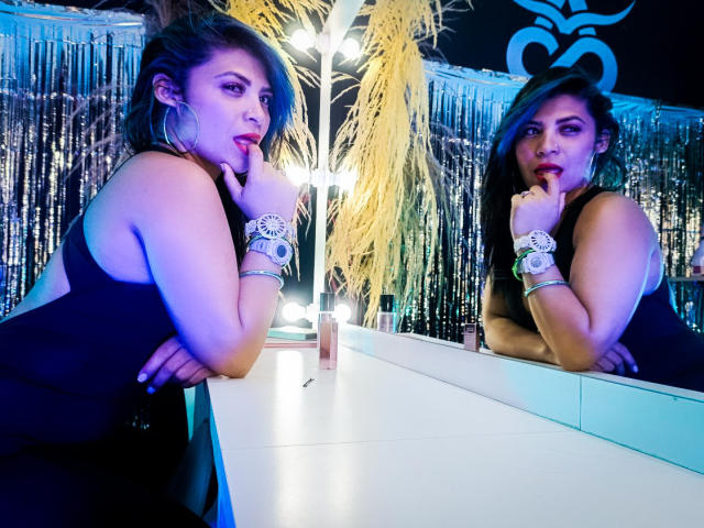 KatalinaMachado with her finger in her mouth, wearing a black tank top, silver bracelet, and wristwatch, reflected in a mirror, with blue and yellow lighting.