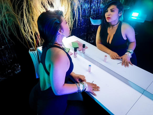 KatalinaMachado with large tits, black dress, and red lipstick, applies makeup in a brightly lit mirror, her hand on a white countertop with makeup items.