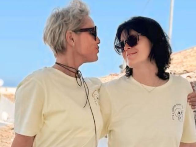 Un couple, l'un avec des cheveux gris et des lunettes de soleil, tenant le bras d'un autre couple avec des cheveux noirs et des lunettes de soleil, tous deux portant des chemises crème, souriant l'un à l'autre, par une journée ensoleillée..