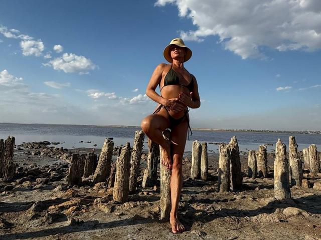 PleasureWeCouple stands on rocky beach, wearing a green bikini, hat, and sun, with one leg raised, showing her toned ass and small tits, against a cloudy sky.