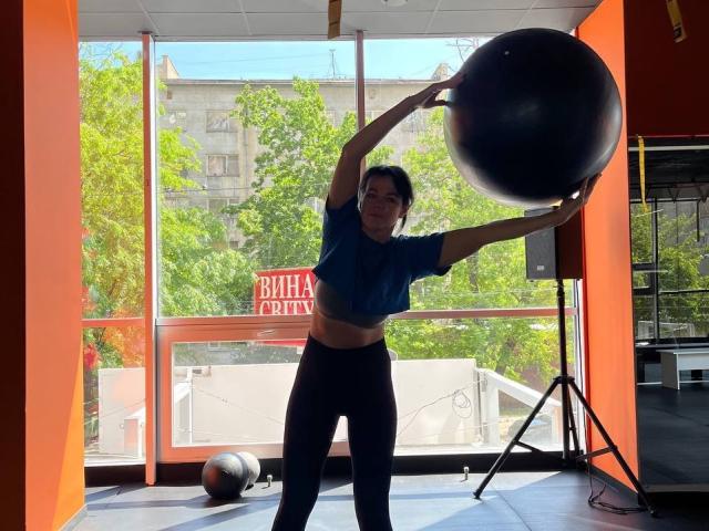 PleasureWeCouple, con cabello oscuro, busto medio, vistiendo una camiseta corta azul y pantalones negros, sostiene una gran pelota de ejercicio negra en un gimnasio luminoso con paredes de color naranja..