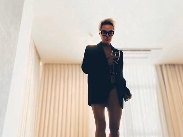 PleasureWeCouple está em um quarto, vestindo uma jaqueta preta, segurando um telefone, usando óculos, com cabelo loiro curto e uma expressão confiante, mostrando sua bunda à mostra..