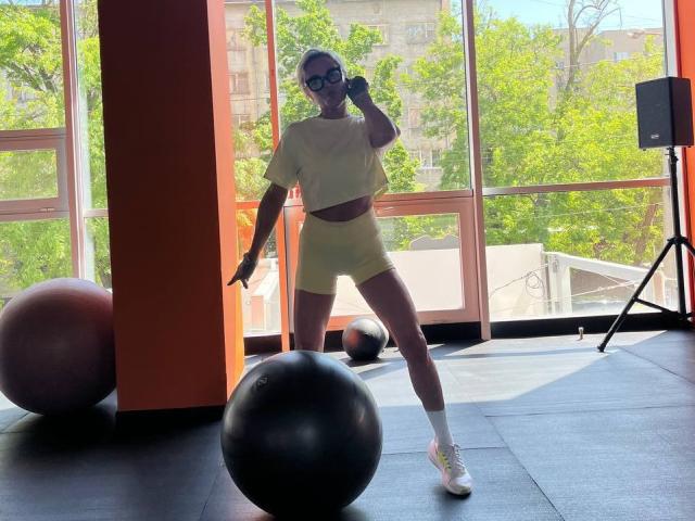 PleasureWeCouple, in yellow crop top and shorts, standing with one leg raised, holding a phone to her ear, in a sunlit gym with exercise balls and a tripod.