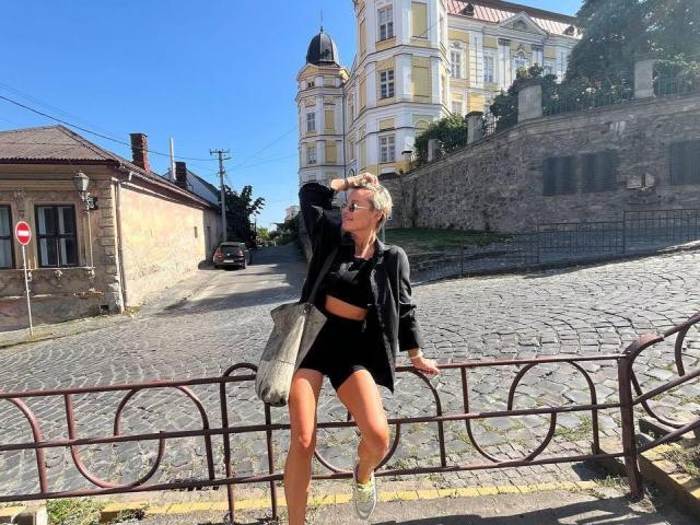 PleasureWeCouple with blonde hair, black crop top, black shorts, and black jacket, sitting on a metal fence, holding a gray bag, under bright blue sky.