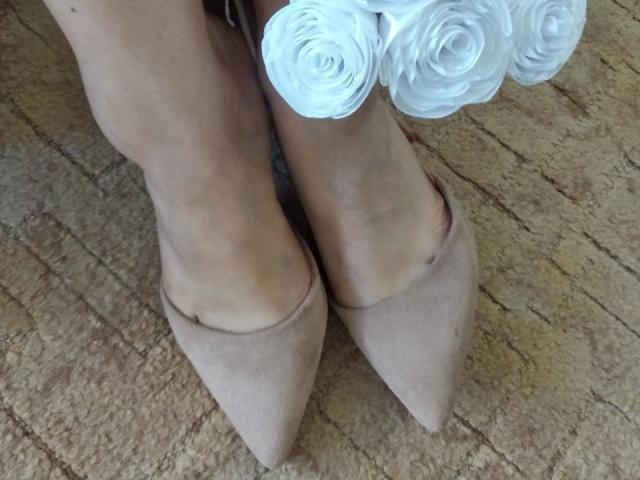 Ses pieds, chaussés d'escarpins pointus en daim beige, ornés de pétales de rose blanche, reposent sur un tapis beige texturé. Ses chevilles sont légèrement croisées..