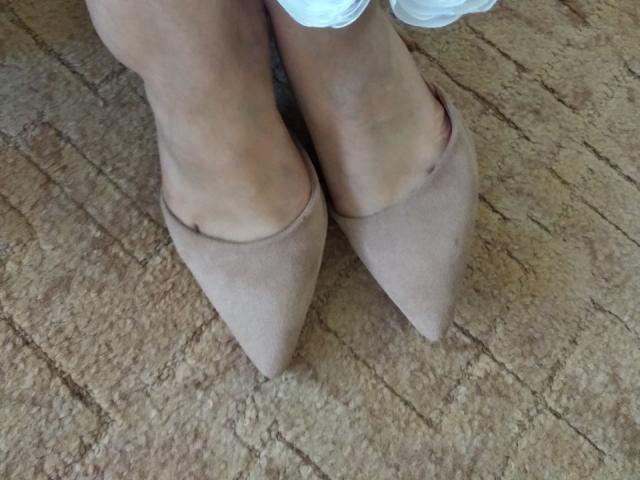 I piedi di Nurri, calzati di eleganti scarpe beige in camoscio con la punta leggermente visibile, sono appoggiati su un tappeto marrone con una texture particolare. Le sue gambe sono leggermente incrociate alla caviglia..