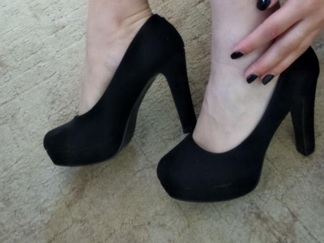 As pernas pálidas de Nurri, calçadas com sapatos de salto alto pretos, exibindo unhas pintadas de preto, estão sobre um tapete texturizado. Sua mão direita repousa sobre o tornozelo..