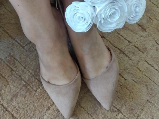 Los pies de Nurri, calzados con tacones puntiagudos de gamuza color beige, adornados con un ramo de rosas blancas, están sobre una alfombra beige con textura. Sus piernas están ligeramente cruzadas..