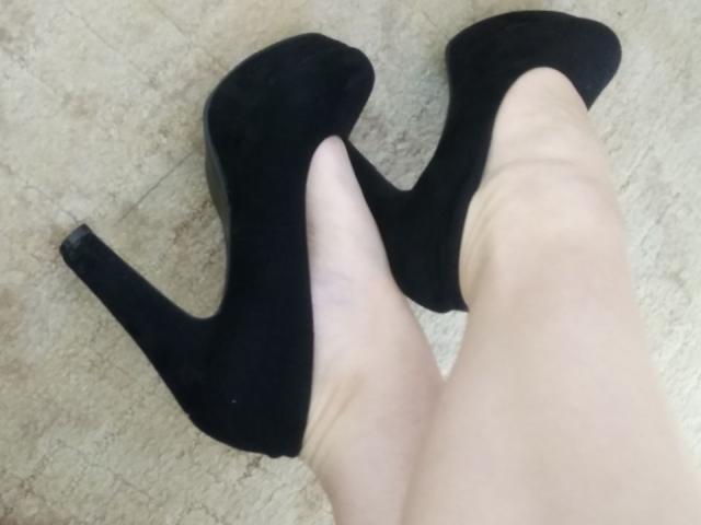 Nurris blasse Beine in schwarzen High Heels, wobei die Absätze nach oben zeigen und die Ausschnitte an den Seiten der Schuhe sichtbar sind. Ihre Füße stehen leicht auseinander auf einem beige farbenen Boden..