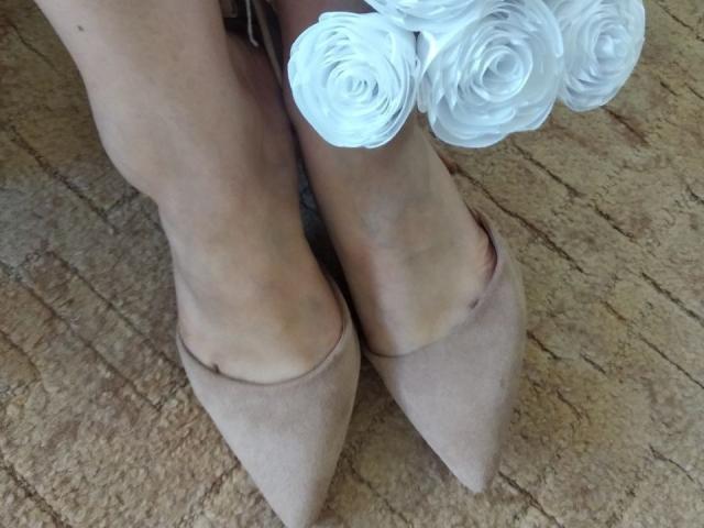 Nurris Zehen stecken in spitzen, beigen Schuhen, mit einem weißen Rosenstrauß an ihren Füßen, und sie steht auf einem strukturierten, beigen Teppich. Ihre Haut ist hell..