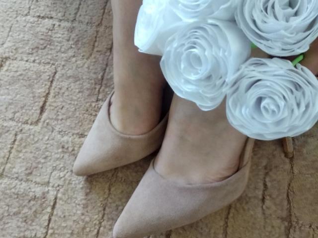 Nurri's feet in beige pointy-toe flats with white rose bouquet on top, showing her pale skin and subtle ankle lines.