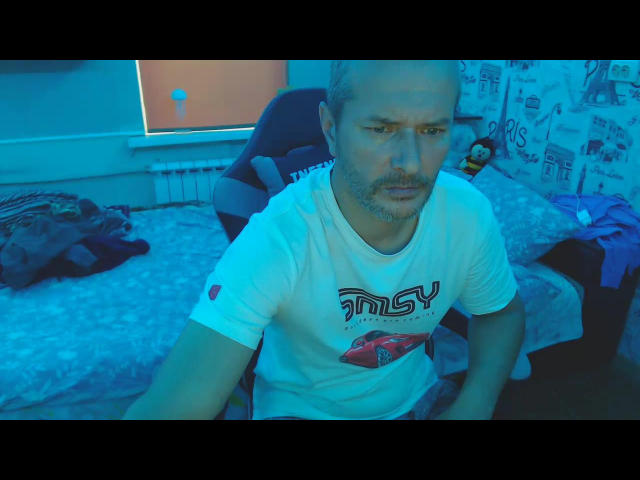 JasperSexy hat einen glatzköpfigen Kopf, einen kurzen Bart und ein ernstes Gesicht. Er trägt ein weißes Hemd mit dem Aufdruck "MS" und einer Grafik eines roten Autos und sitzt in einem blau beleuchteten Raum..