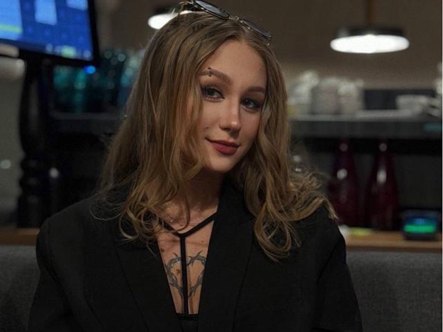 Alice, com cabelo loiro ondulado, blazer preto e tatuagens no peito, usando batom vermelho, com um leve sorriso, óculos na cabeça, sentada em um escritório com pouca luz..