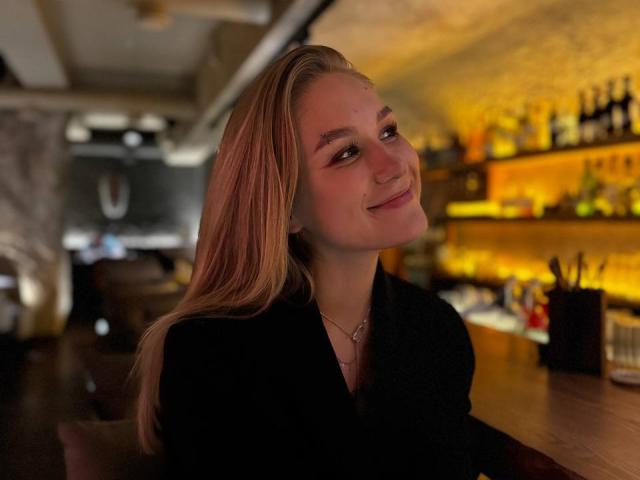 AliceVibes with long blonde hair, smiling, wearing a black top, sitting at a bar, warm lighting, soft expression, subtle makeup, slight tilt of her head, necklace visible.